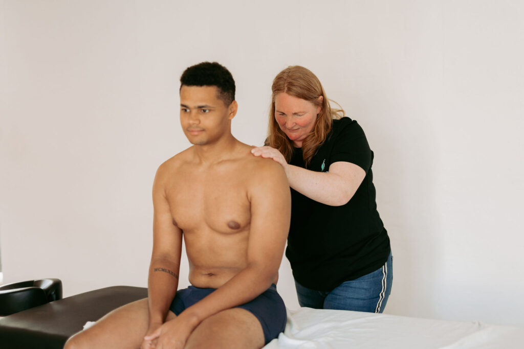 Professionel massør behandler spændinger i skulder og nakke på en siddende klient i forbindelse med massage i eget hjem.